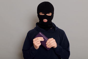 Serious young man burglar in robbery mask and black sweater holds tightly wallet that he just...