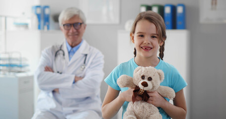 Smiling healthy little girl hugging teddy bear in doctor office looking at camera.