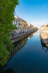 Fototapeta premium Perspective of the famous old Navigli neighborhood in Milan, Italy. Water canal in the foreground, looking like a river, with a green tree. Buildings and blue sky in the background.
