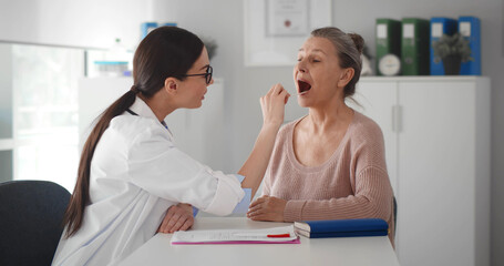 Female doctor checking throat of senior patient while examining her during consultation in clinic