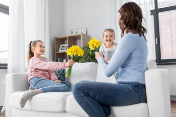 people, family and holidays concept - two daughters giving daffodil flowers to happy mother at home