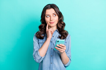 Photo of excited thoughtful lady dressed denim shirt smiling holding modern gadget looking empty space isolated teal color background