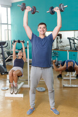 Cheerful sporty guy doing exercises with dumbbells at gym