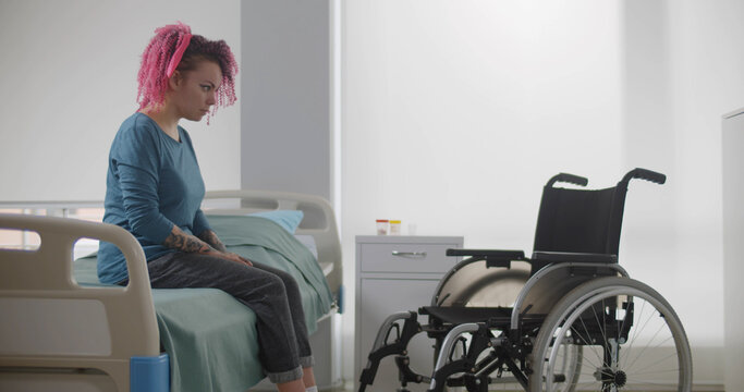 Young woman patient sitting on hospital bed looking at wheelchair 