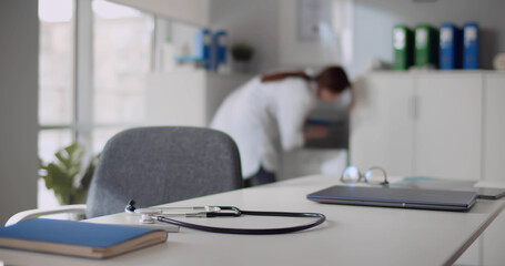 Stethoscope and documents lying on desk in clinic office with medical staff walking on background
