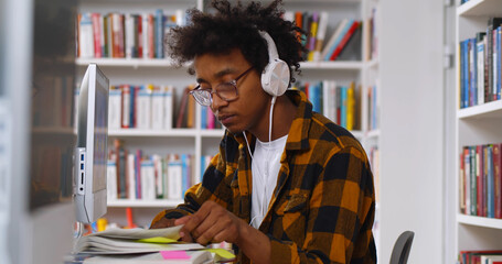 African student in headphones learning online at computer sitting in library in university.