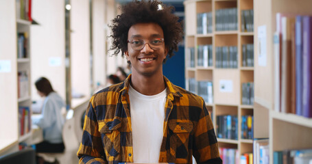 Confident handsome african student smiling at camera with library bookshelves on background
