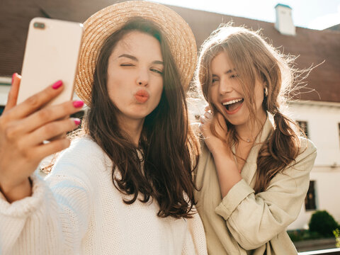 Two young beautiful smiling hipster female in trendy white sweater and coat. Sexy carefree women posing on the street background in hats. Positive models having fun outdoors. Hugging and taking selfie