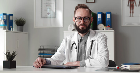 Fototapeta premium Portrait of handsome medical worker smiling at camera in hospital office wearing white coat