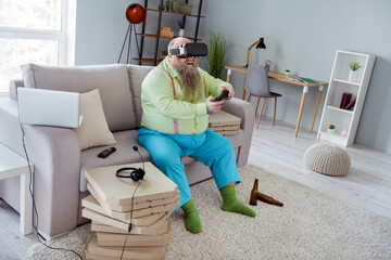 Photo of fat impressed big belly man wear green shirt vr eyewear sitting sofa holding playstation indoors house room