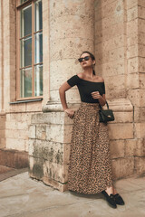 Fototapeta na wymiar Young pretty beautiful tanned caucasian woman with pony tail brunette hair in black top and leopard skirt standing leaning on old palace building. Sunny summer day