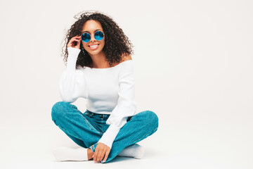 Beautiful black woman with afro curls hairstyle.Smiling model in sweater and trendy jeans clothes. Sexy carefree female posing on white background in studio. Tanned and cheerful. In sunglasses