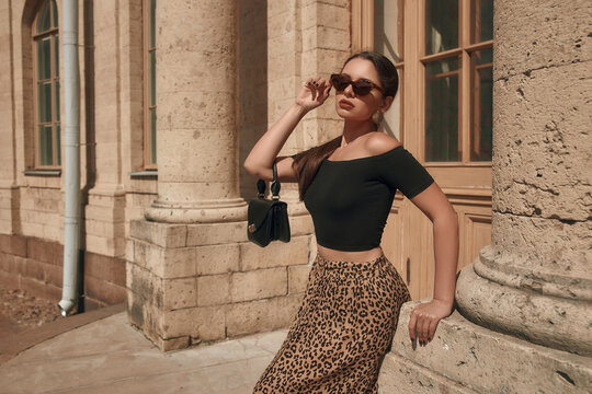 Young pretty beautiful tanned caucasian woman with pony tail brunette hair in black top and leopard skirt standing leaning on old palace building. Sunny summer day