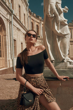 Young beautiful sexy tanned caucasian woman with perfect mak-up and pony tail brunette hair in black top and leopard skirt standing against old palace on a sunny day