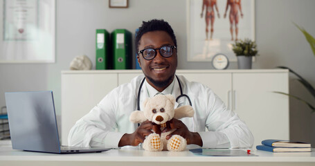 Portrait of african young pediatrician sitting at desk with teddy bear smiling at camera