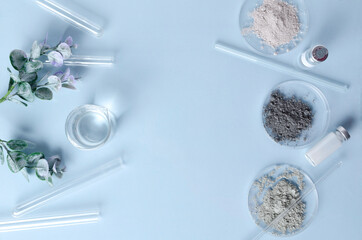 Top view of lab desk and equipment for beauty product testing on it.Glass tubes,sticks, dishes, facial dry powder clay and green leaves on the table.Empty space