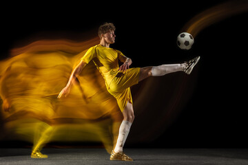 Young caucasian male football or soccer player kicking ball for the goal in mixed light on dark background. Concept of healthy lifestyle, professional sport, hobby.