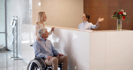 Professional receptionist talking to senior woman and handicapped man at desk in clinic