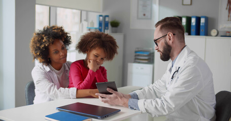 Young male doctor showing medical test results on tablet to young woman with little daughter