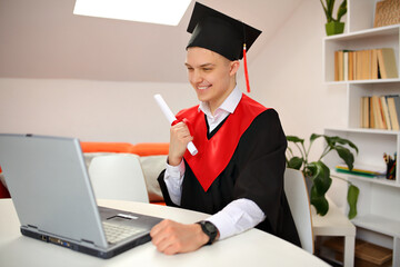 A student in graduation gown and a graduate cap watches the broadcast of the graduation ceremony while at home