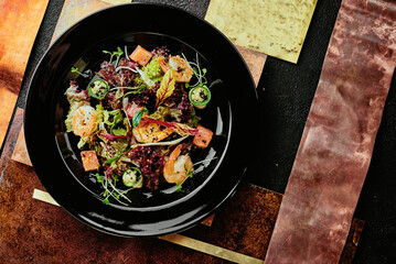 Salad with salmon, shrimps, lettuce, cucumber and soybean sprouts in a black plate on a background of copper plates.
