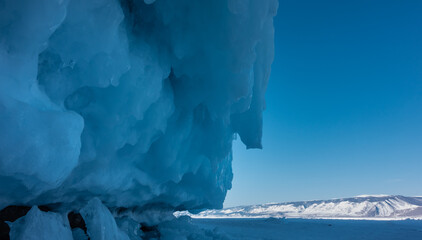The base of the rock is covered with a thick layer of ice splashes and bizarre icicles. Close-up. A mountain range covered with snow is visible in the distance. Lake Baikal in winter.
