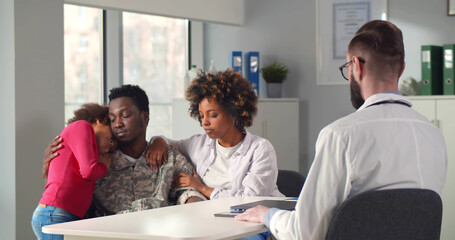 Sad african wife and little daughter supporting young soldier in doctor office