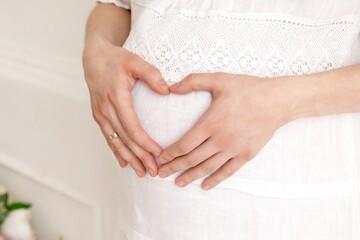 One pregnant woman in a white dress with a wedding ring holds her hands on her stomach while in a bright clean room