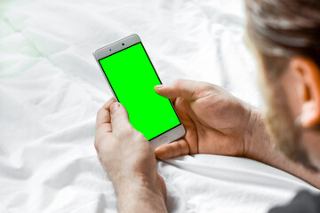 Young Bearded Man Using Green Chroma Key Screen Phone, Relaxing in a Bed. Guy Uses Green Mock-up Screen Smartphone. Rear View. Man lying on white blanket and Watching Video on ChromaKey display