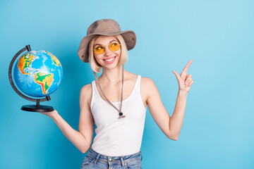 Fototapeta premium Portrait of charming cheerful girl hand hold globe look indicate finger empty space isolated on blue color background