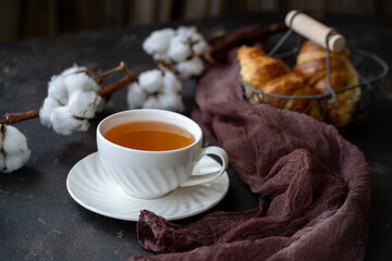 Tea in the morning. The white cup is on a white saucer. Hot tea is poured into the cup. On a dark background next to the cup is a metal basket with two croissants. There is a cotton twig at the back