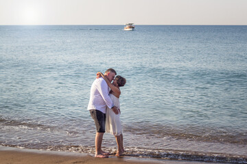 Ein Pärchen steht am Strand umarmen sich und küsst sich. Im Hintergrund ist Meer und Schiff. Es sind viele Wellen. Die Frau hat ein weißes Kleid an, der Mann ein weißes Hemd. Sie genießen den Urlaub