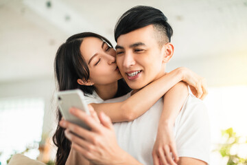  Happy young couple kissing and  looking at mobile phone in living room at home