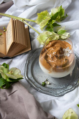 Delicious dalonga coffee with ice and milk in glass cup. Iced frothy Dalgona Coffee, a trendy fluffy creamy whipped coffee. Korean drink latte espresso with coffee foam. Green flowers and a book.