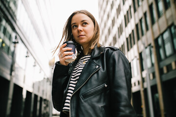 Young pretty girl listening to music in the big city. girl in a leather jacket with headphones drinking coffee near business center . smiling and loathing