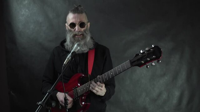 Bearded rocker with shaved temples in black hoodie and round glasses plays red electric guitar, sings stage on nightclub concert with black curtain background