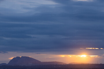 A Panoramic Landscape Silhouette of hills at the bottom with dark clouds in the horizon with sun peeping through them
