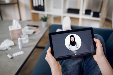 Tablet screen view, close up. Top view of unrecognizable female patient, sitting with digital tablet pc on couch and making video call to young professional Arabian doctor from home.