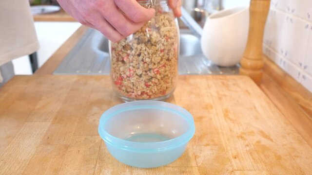 Pouring roasted oatmeal from a glass into a blue plastic bowl. Wooden table in the kitchen