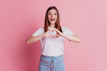 Cheerful young beautiful girl is gesturing a heart with fingers excited face look camera on pink wall