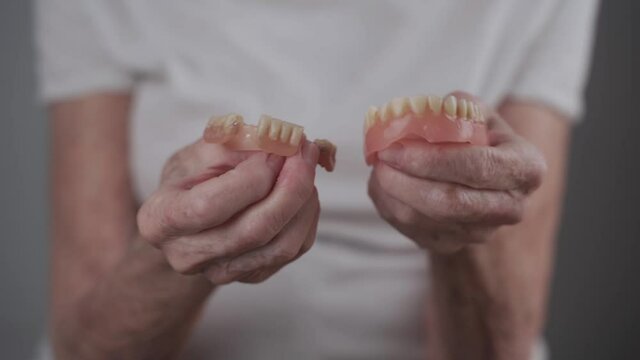 Senior woman holding close-up of dental prosthesis in hands. Old female shows false teeth. Removable denture in hands of an elderly patient in studio on gray background.
