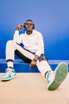 Young afro American black man wearing a white sweatshirt, basketball sneakers and headphones seated on a blue wall looking at camera with confidence