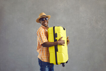 Cheerful man going on summer vacation. Portrait of happy smiling black tourist in glasses, sun hat and orange shirt holding yellow travel suitcase standing on gray studio background