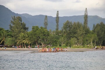 A group of colorfully dressed Vanuatans congregating on a beach near Mele, Port Vila, for a Sunday stroll, jungle covered mountains and a cloudy sky in the background
