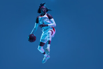 Beautiful african-american female basketball player in motion and action in neon light on blue background. Concept of healthy lifestyle, professional sport, hobby.