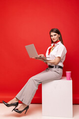 A young woman in a white shirt and trousers holds a laptop and a coffee cup