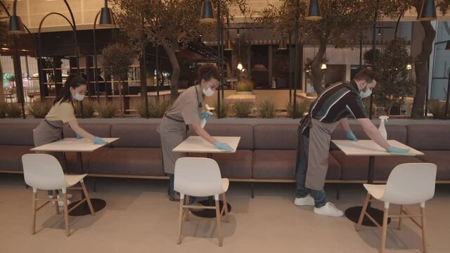 Wide shot of three diverse multiethnic restaurant workers wearing face masks, gloves and aprons, disinfecting tables, wiping them with towels, standing in row