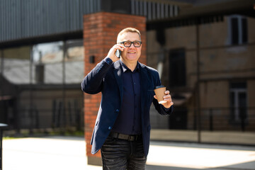 Businessman talking by smartphone and smiling at camera. Cheerful middle aged businessman holding disposable coffee cup and talking by cell phone on street. Communication concept