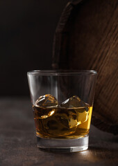 Glass with ice cubes of scotch whiskey next to wooden barrel and black.