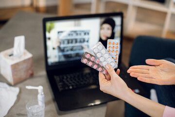 Medicine, online appointment concept. Hands of patient, sitting on sofa at home with blisters pills in hand, having video chat with female Muslim doctor on laptop and consulting about treatment.